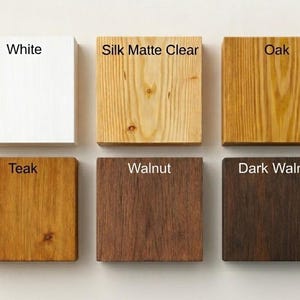 Op de afbeelding: Zes vierkante houten stalen gerangschikt in twee rijen. De bovenste rij bevat de afwerkingen White, Silk Matte Clear en Oak. De onderste rij toont Teak, Walnut en Dark Walnut. Elk staal is gelabeld met de bijbehorende kleurnaam.