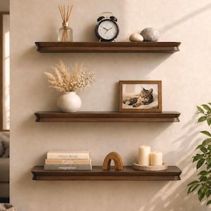Op de afbeelding: Drie donkerbruine houten zwevende planken gemonteerd aan een muur. De bovenste plank bevat een geurstokjeshouder, een zwarte wekker en twee stenen. De middelste plank toont een witte vaas met gedroogde planten en een ingelijste foto van een kat. De onderste plank bevat drie gestapelde boeken, een houten boog en twee kaarsen op een schaal.