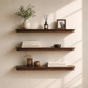 May include: Three horizontal dark brown wooden floating shelves mounted on a light beige wall. The top shelf holds a white vase with greenery, a reed diffuser, and a small framed cat photo. The middle shelf holds three books and a small dark bowl.