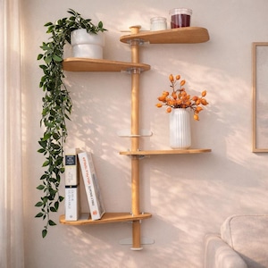 May include: A wall-mounted shelving unit featuring a central vertical wooden pole with four asymmetrical, rounded light wood shelves. The shelves hold a white pot with a trailing green plant, two glass jars, a white vase with orange berries, and two books.