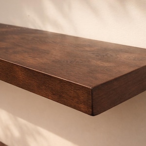Pode incluir: Uma prateleira de madeira castanha escura, retangular, montada numa parede clara. A prateleira tem uma superfície lisa e polida e um tom rico e quente. O grão da madeira é visível, adicionando textura. Design simples e moderno.