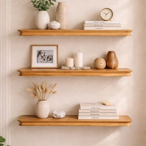 May include: Three horizontal wooden wall shelves featuring rounded edges. The top shelf holds a ribbed white vase with greenery, a beige vase, and a stack of books with a gold clock. The middle shelf displays a framed dog photo, two white candles, and two brown vases. The bottom shelf holds a ribbed white vase with dried grass, a white stone, and a stack of books.