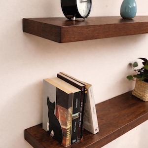 Può includere: Due mensole galleggianti in legno marrone scuro su una parete bianca. La mensola superiore contiene una sveglia nera e un piccolo vaso blu con fiori bianchi. La mensola inferiore espone libri e un fermalibri a forma di gatto nero.