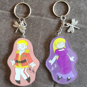 Puede incluir: Dos llaveros con personajes de dibujos animados. El llavero de la izquierda presenta a un hombre con un uniforme rojo y dorado con una gema azul en la manga. El llavero de la derecha presenta a una mujer con un vestido morado y guantes blancos. Ambos llaveros tienen un dije de lib&eacute;lula plateado.