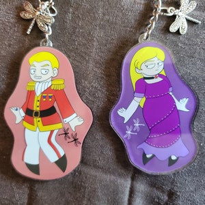 Puede incluir: Dos llaveros de personajes coloridos con un dije de lib&eacute;lula. El llavero de la izquierda presenta un personaje masculino con un uniforme rojo y dorado con una gema azul en la manga. El llavero de la derecha presenta un personaje femenino con un vestido morado y guantes blancos. Ambos llaveros tienen un fondo rosa y morado con detalles de lib&eacute;lula.