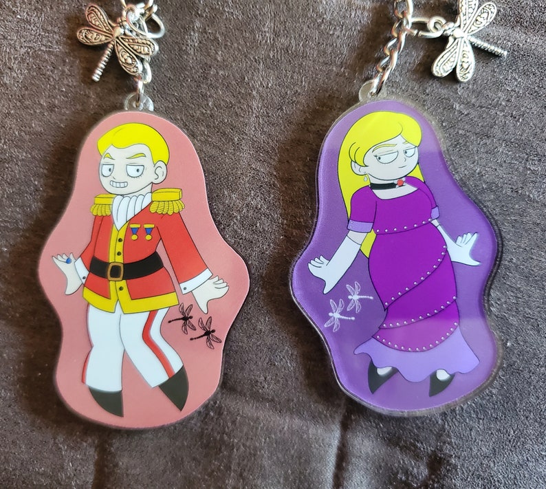 Puede incluir: Dos llaveros de personajes coloridos con un dije de lib&eacute;lula. El llavero de la izquierda presenta un personaje masculino con un uniforme rojo y dorado con una gema azul en la manga. El llavero de la derecha presenta un personaje femenino con un vestido morado y guantes blancos. Ambos llaveros tienen un fondo rosa y morado con detalles de lib&eacute;lula.