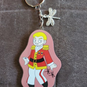 Puede incluir: Un llavero rosa con un personaje de dibujos animados que lleva un uniforme rojo y dorado con pantalones blancos y zapatos negros. El personaje tiene el pelo rubio y una gema azul en el dedo. Dos lib&eacute;lulas negras est&aacute;n en el llavero.