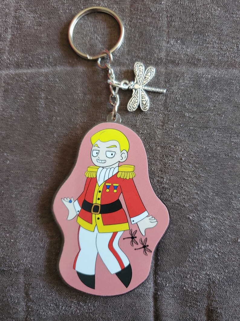 Puede incluir: Un llavero rosa con un personaje de dibujos animados que lleva un uniforme rojo y dorado con pantalones blancos y zapatos negros. El personaje tiene el pelo rubio y una gema azul en el dedo. Dos lib&eacute;lulas negras est&aacute;n en el llavero.