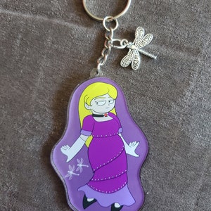 Puede incluir: Un llavero con un personaje de dibujos animados morado y blanco con un amuleto de lib&eacute;lula plateado. El personaje es una mujer con cabello rubio que lleva un vestido morado y guantes blancos.