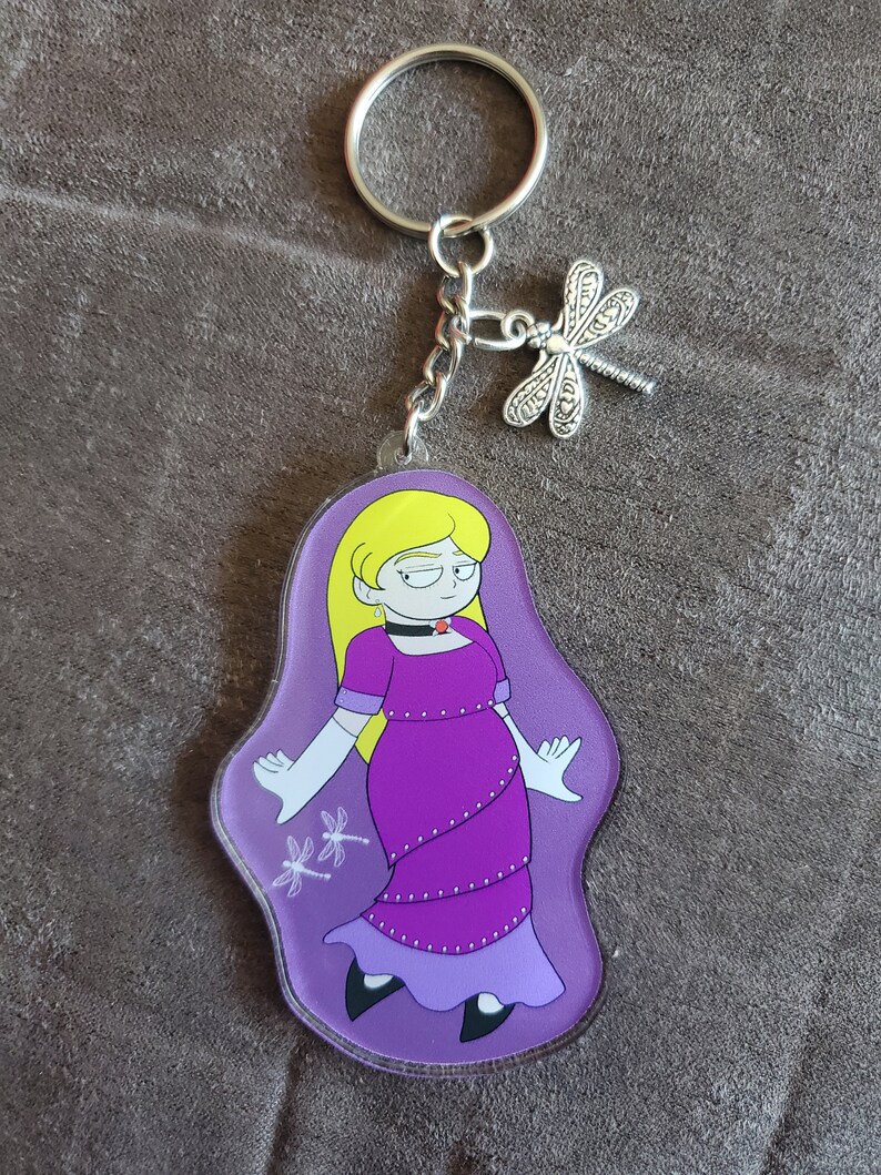 Puede incluir: Un llavero con un personaje de dibujos animados morado y blanco con un amuleto de lib&eacute;lula plateado. El personaje es una mujer con cabello rubio que lleva un vestido morado y guantes blancos.