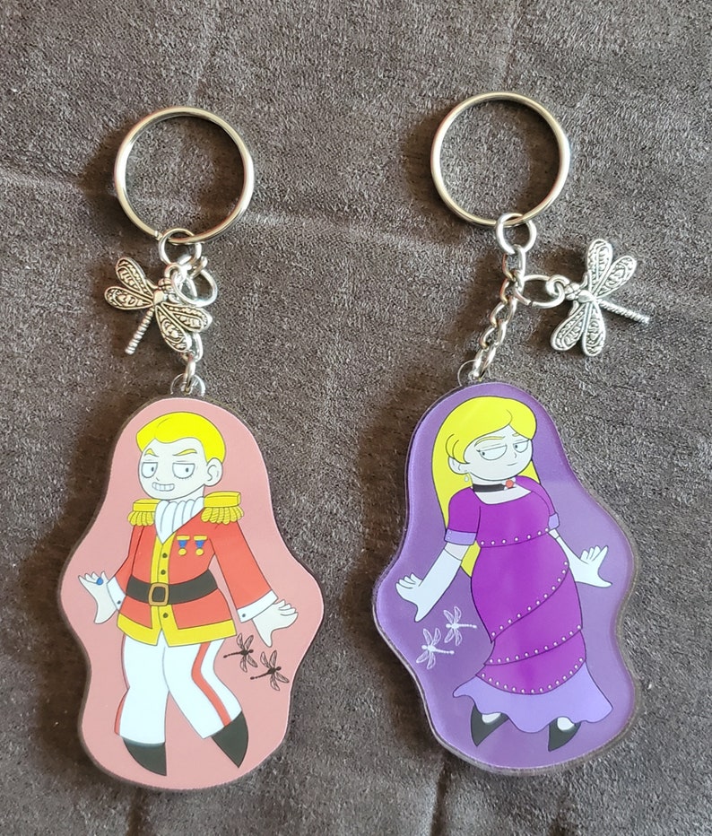 Puede incluir: Dos llaveros con personajes de dibujos animados. El llavero de la izquierda presenta a un hombre con un uniforme rojo y dorado con una gema azul en la manga. El llavero de la derecha presenta a una mujer con un vestido morado y guantes blancos. Ambos llaveros tienen un dije de lib&eacute;lula plateado.