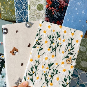 May include: A collection of rectangular fabric-covered journals featuring intricate embroidery designs. One journal displays white daisy flowers with yellow centres, while another features small cat faces and paw prints on a cream background.