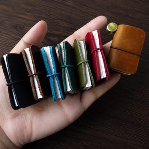 Op de afbeelding: Een verzameling van zeven miniatuur notitieboekjes met een leren kaft in diverse kleuren zoals zwart, bruin, blauw, groen en rood. Elk klein notitieboekje heeft een elastische sluiting en witte pagina's, gepresenteerd in de palm van een hand.