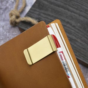 Puede incluir: Un clip metálico de color dorado para bolígrafo sujeto a una cubierta de cuaderno de cuero color canela. Un bolígrafo blanco con detalles rojos y el texto AIHAO está asegurado en el cuaderno de páginas color crema. El clip tiene un acabado.