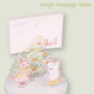 May include: Acrylic message holder featuring a clear base with a postcard and cartoon characters. The postcard has a stamp and lines. The characters include a pink-dressed figure, a white figure, and a decorated Christmas tree. The text "Acrylic message holder" is at the top.