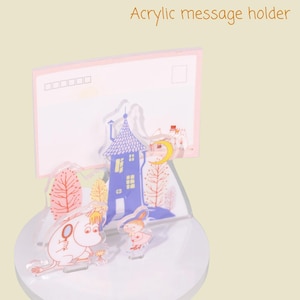 May include: Acrylic message holder featuring a whimsical design with cartoon characters, a blue house, and trees. The holder has a clear base and a pink postcard-shaped back. The text "Acrylic message holder" is visible.
