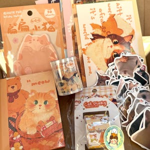 May include: A selection of cat-themed stationery, including sticky notes, stickers, a notebook, and decorative tape. The items feature illustrations of cats in various poses and settings, with a warm colour palette of orange, peach, and brown.