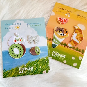 Op de afbeelding: Twee kaarten met geborduurde patches met voedsel- en natuurthema's. Eén kaart heeft een kiwi, een strik en een hondachtig karakter. De andere kaart heeft een sinaasappelschijfje, ijs en een croissant. De kaarten lezen "Embroidery Patch" en "fluffstuff".