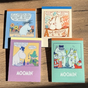 May include: Four Moomin-themed notepads with colorful covers. Each notepad features a different illustration of Moomin characters with text. The notepads have blue, orange, yellow, and teal borders. The text includes phrases like "Isn't it wonderful" and "Our own house."