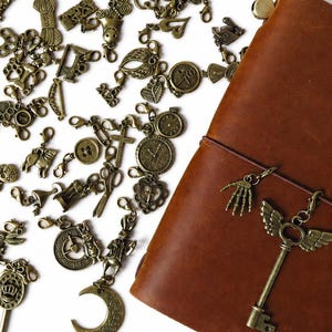 May include: A collection of assorted antique bronze-toned metal charms featuring various shapes including keys, moons, clocks, and animals, scattered next to a rectangular brown leather-bound notebook with a winged key charm attached to its elastic band.