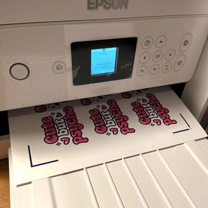 May include: A white Epson printer with a digital display screen showing a printing status message. A sheet of white paper is emerging from the printer, featuring multiple repeating pink and black graphic designs that read "Cute but psycho".