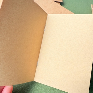 May include: An open notebook with light brown, unlined paper pages. The booklet is held together at the center fold by two silver metal staples. The texture of the paper is matte and fibrous, showing a natural tan color throughout the interior.
