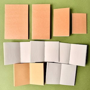 Op de afbeelding: Een verzameling notitieboekjes met effen beige kaften in verschillende formaten, gerangschikt naast open notitieboekjes met verschillende stijlen binnenpagina's, waaronder horizontale lijnen, stippenrasters, blanco pagina's en ruitpatronen.
