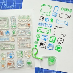 May include: Clear acrylic stamps with travel-themed designs, including trees, a train, and the word "HAPPY!". The stamps are in green, blue, and orange. A sheet of stamped images shows the designs in use, with text like "New York" and "Travel plan".
