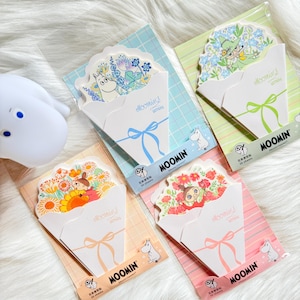 Op de afbeelding: Vier Moomin-thema briefpapier sets. Elke set heeft een wit papieren boeket ontwerp met een kleurrijk bloemstuk en een Moomin-personage. De sets zijn verpakt op pastelkleurige achtergrondkaarten met de woorden "MOOMIN" en "LETTERS" erop gedrukt.