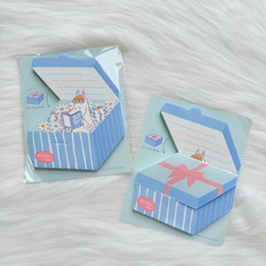 May include: Two blue and white striped gift box-shaped sticky note pads. One box is open, revealing a cartoon character reading a book. The other box is closed with a pink bow. The background is a light blue card.