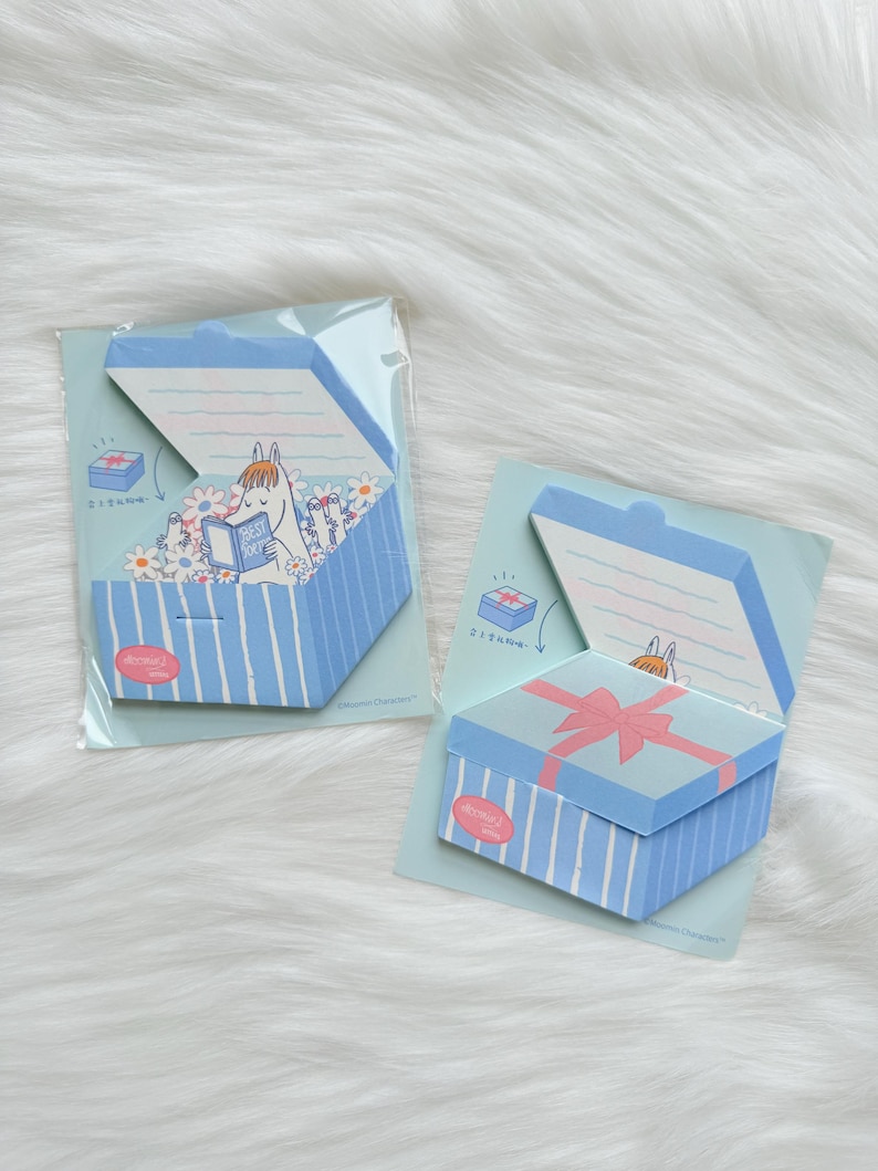 May include: Two blue and white striped gift box-shaped sticky note pads. One box is open, revealing a cartoon character reading a book. The other box is closed with a pink bow. The background is a light blue card.