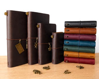 May include: A collection of leather journals in various sizes and colors. Four dark brown journals stand upright with brass charms, while a stack of seven journals in black, brown, maroon, blue, green, tan, and red lies horizontally to the right.