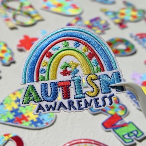 May include: An embroidered patch featuring a rainbow arch with puzzle piece patterns in red, yellow, green, and blue. The text "AUTISM AWARENESS" is stitched in bold, multi-coloured letters below the rainbow on a white fabric background.