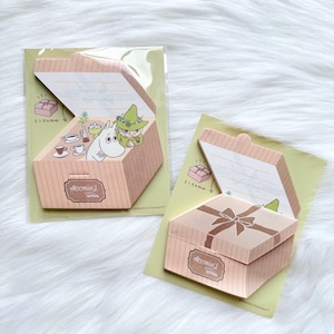 May include: Two decorative paper boxes with a light brown color and the text "Moomin's Letters". One box is open, revealing a cartoon illustration of a character in a green hat. The other box is closed with a bow on top. Both boxes are on a light green background.