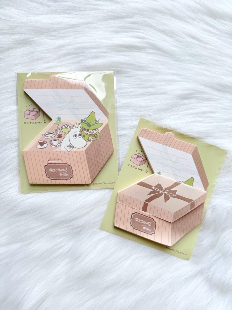 May include: Two decorative paper boxes with a light brown color and the text "Moomin's Letters". One box is open, revealing a cartoon illustration of a character in a green hat. The other box is closed with a bow on top. Both boxes are on a light green background.