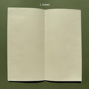 May include: An open notebook with off-white pages featuring a light gray dotted grid pattern. The center fold is secured with visible white thread stitching. The text 1. Dotted is printed in white at the top center of the notebook pages.