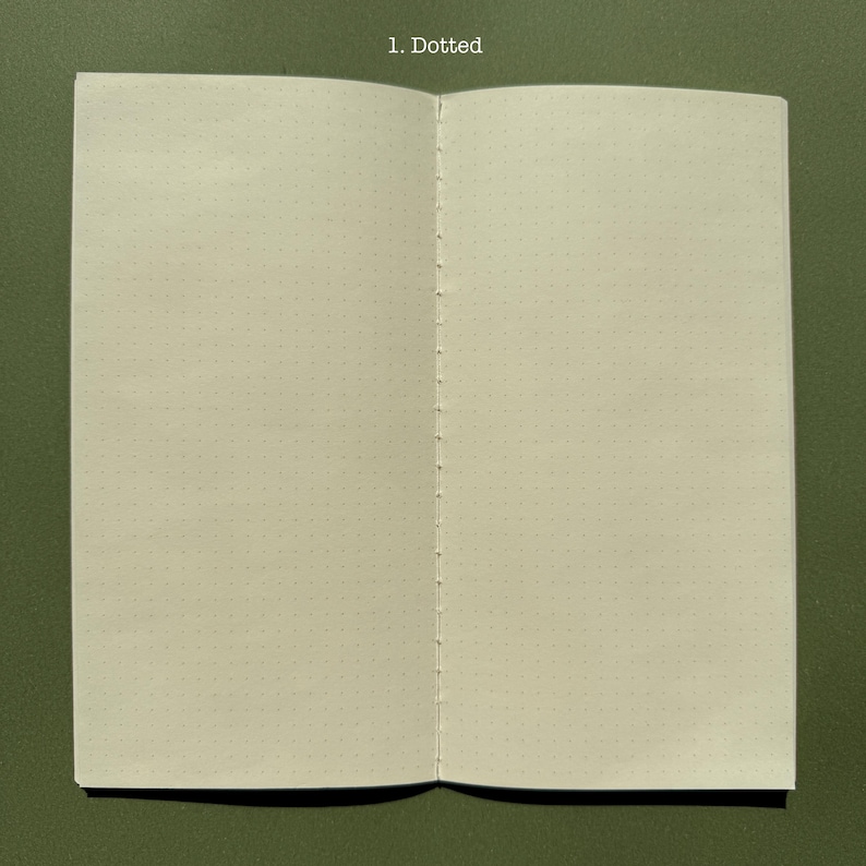 May include: An open notebook with off-white pages featuring a light gray dotted grid pattern. The center fold is secured with visible white thread stitching. The text 1. Dotted is printed in white at the top center of the notebook pages.