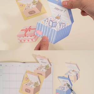 May include: A collection of decorative paper boxes with pop-up designs. One box is blue with white stripes and the word "surprise" on the lid. Another is pink with a bow design. The boxes feature cartoon characters and floral elements.