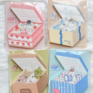 May include: Four Moomin-themed sticky note pads in the shape of open boxes. Each pad features a different design: a pink cake, a yellow box with stars, a brown box with tea, and a blue box with flowers. Each box has a Moomin character inside.