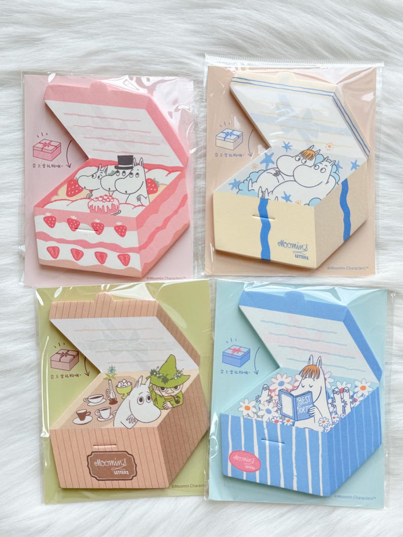 May include: Four Moomin-themed sticky note pads in the shape of open boxes. Each pad features a different design: a pink cake, a yellow box with stars, a brown box with tea, and a blue box with flowers. Each box has a Moomin character inside.