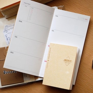 Pode incluir: Um caderno de planeamento semanal aberto com páginas brancas apresentando um layout Plan of Week de segunda a domingo, emparelhado com um livreto de inserção bege menor Traveler's Notebook Weekly com o texto Weekly Travel Notes na capa frontal.
