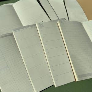 May include: A collection of open planners and notebooks with off-white pages featuring various layouts including weekly calendars, daily schedules, and lined sections. The pages display printed text for days of the week and grid patterns in gray ink.