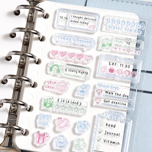 Puede incluir: Pegatinas transparentes para planificador con varios diseños y texto, incluyendo "I bought delicious bread today!", "Today's Record" y "Water Tracker". Otras pegatinas presentan iconos y frases como "skin care", "La La Land" y "Goal". Las pegatinas están en una página blanca.