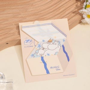May include: A Moomin-themed stationery set featuring a die-cut design. The set includes a light beige card with a blue and white illustration of Moomin characters. The words "Moomins Letters" are printed on the card.