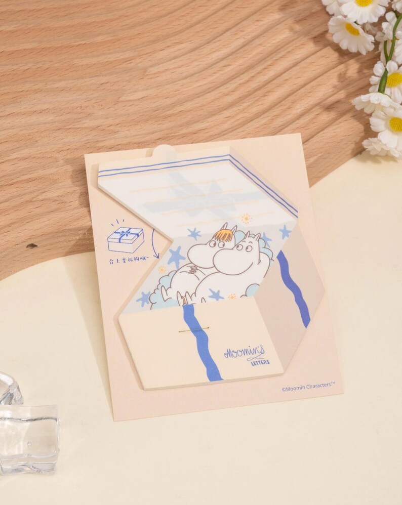 May include: A Moomin-themed stationery set featuring a die-cut design. The set includes a light beige card with a blue and white illustration of Moomin characters. The words "Moomins Letters" are printed on the card.