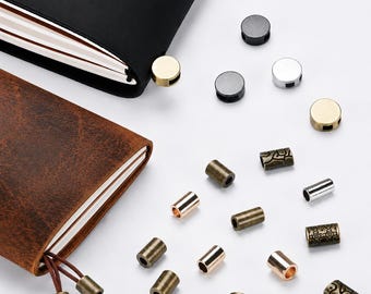 May include: A black leather notebook and a brown distressed leather notebook, each featuring metal charms. Scattered around are various cylindrical and disc-shaped metal beads in silver, gold, bronze, and copper tones, some with etched patterns.