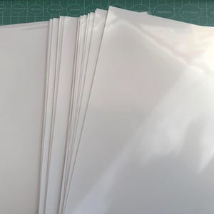 May include: A stack of multiple rectangular sheets of smooth, glossy white paper or cardstock. The sheets are arranged in a slightly fanned-out pile, showing a clean, bright white surface with a reflective finish on the top sheet.