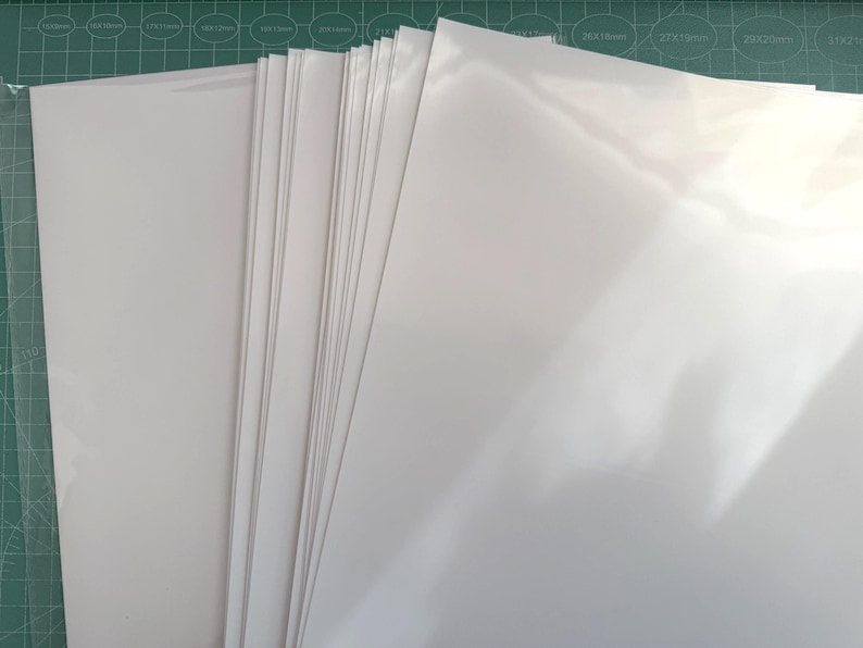 May include: A stack of multiple rectangular sheets of smooth, glossy white paper or cardstock. The sheets are arranged in a slightly fanned-out pile, showing a clean, bright white surface with a reflective finish on the top sheet.