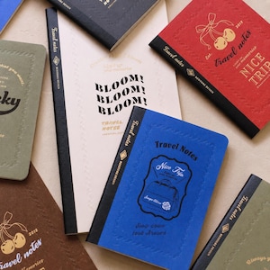 Op de afbeelding: Een verzameling rechthoekige notitieboeken met zwarte ruggen en covers in rood, blauw, olijfgroen, bruin en crème. De covers zijn voorzien van reliëfranden, gouden of zwarte tekst zoals Travel Notes, Nice Trip en Bloom, en grafische illustraties.