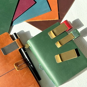 Puede incluir: Un cuaderno de cuero color canela con un bolígrafo negro y un clip de metal plateado, junto a un cuaderno de cuero verde con tres clips de metal dorado. Al fondo, hay varias muestras de cuero superpuestas en verde, morado, azul, naranja y marrón.